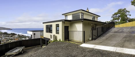A modern two-story house with a view of the ocean, a gravel driveway, and a wooden fence.