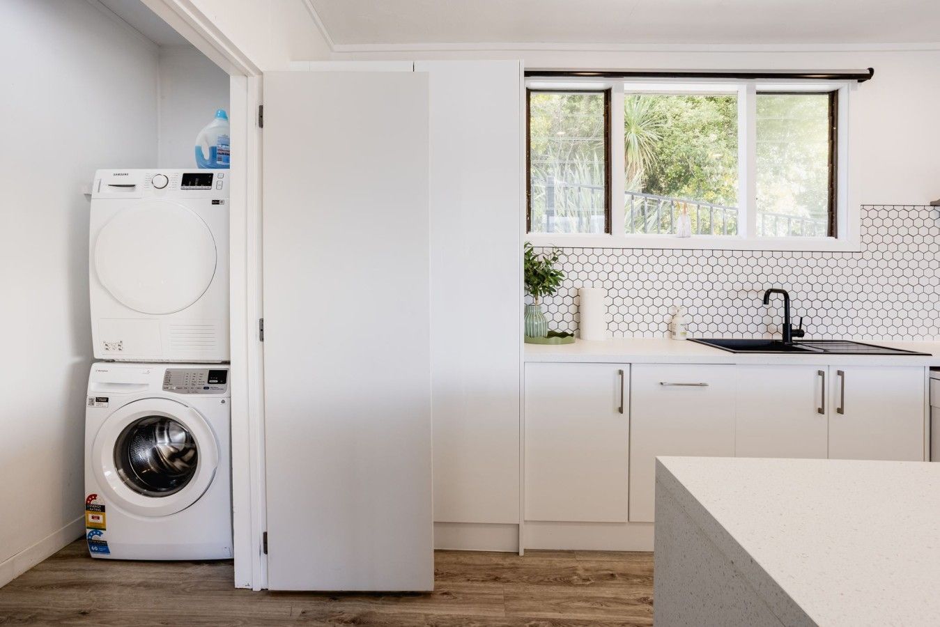 A compact kitchen with a built-in washing machine, a window with a view of greenery, and a tiled backsplash.