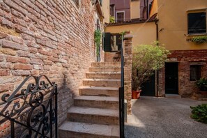 Un estrecho callejón con un muro de ladrillo a un lado y la entrada de un edificio al otro. Se observa un conjunto de escalones de piedra que conducen a la entrada, y una barandilla de metal a la izquierda.