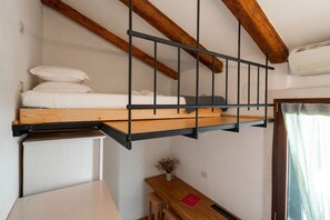 Una cama litera con cabecera de madera y barandillas de metal, una mesita con un florero, y una ventana con cortinas.