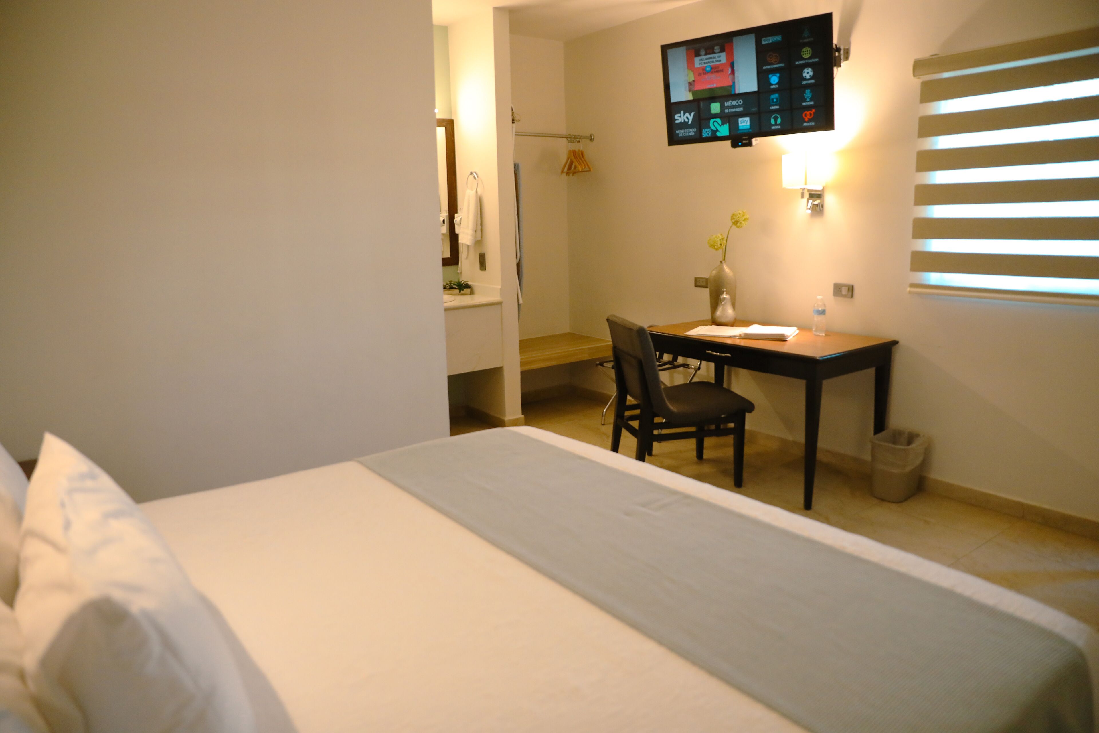 A hotel room with a bed, a desk, a chair, a TV mounted on the wall, and a window with blinds.
