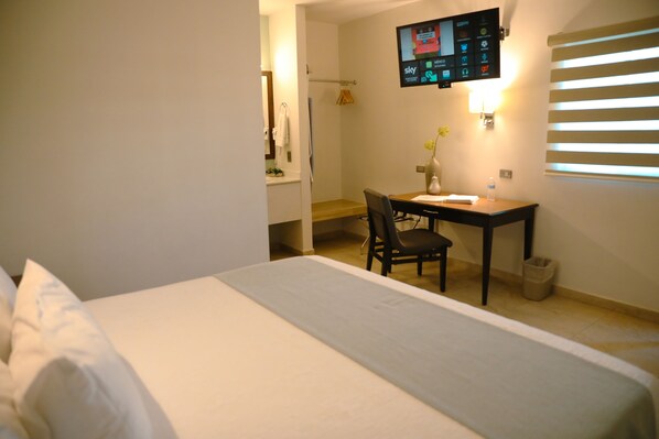 A hotel room with a bed, a desk, a chair, a TV mounted on the wall, and a window with blinds.