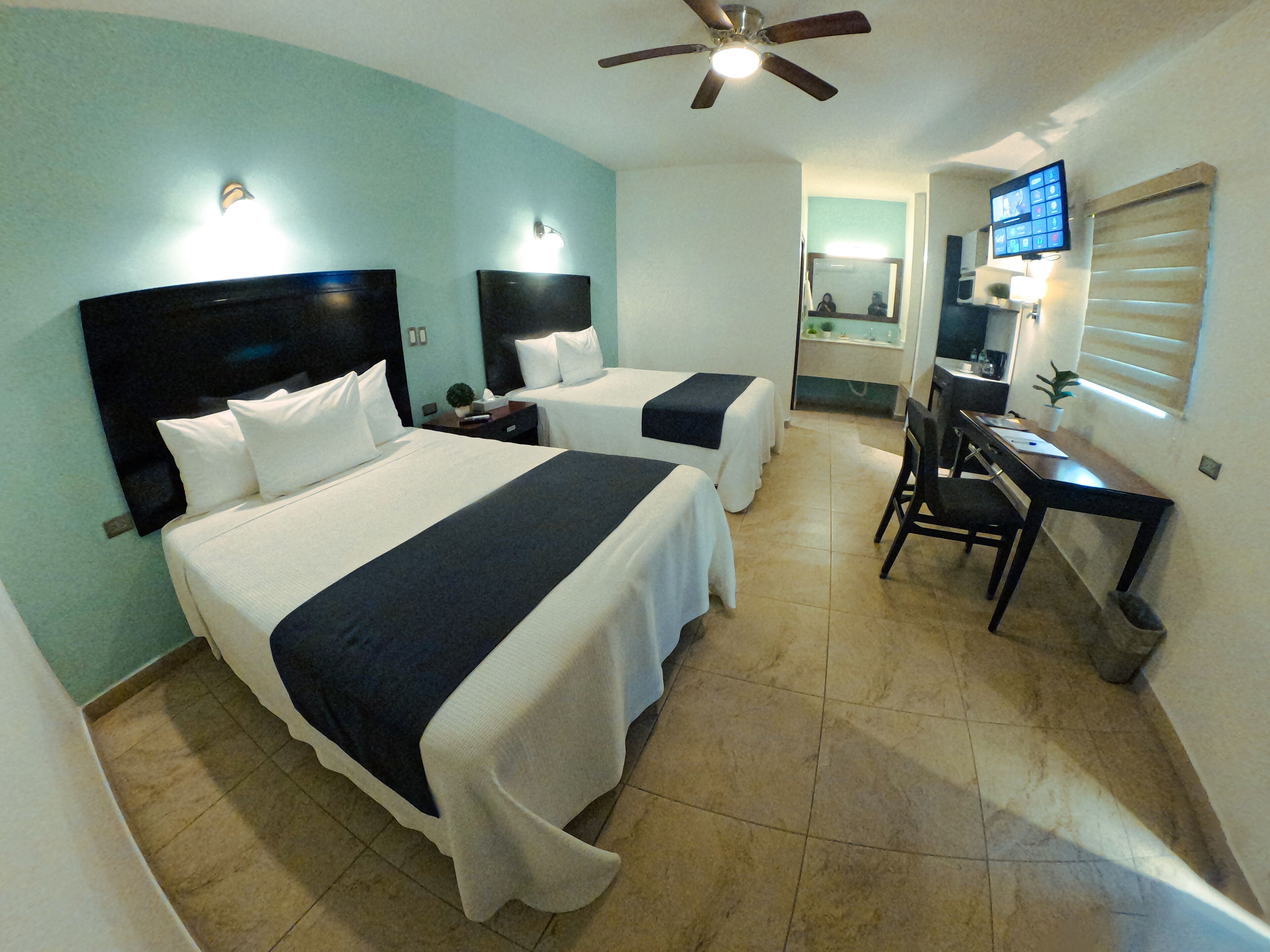 A hotel room with two beds, a desk, a chair, a TV, and a ceiling fan.