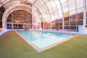 Piscina interior com teto de vidro, rodeada por grama sintética.