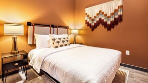 A neatly made bed with a white bedspread and patterned pillows, flanked by two bedside tables with lamps, in a room with a warm wall color and a decorative hanging piece.