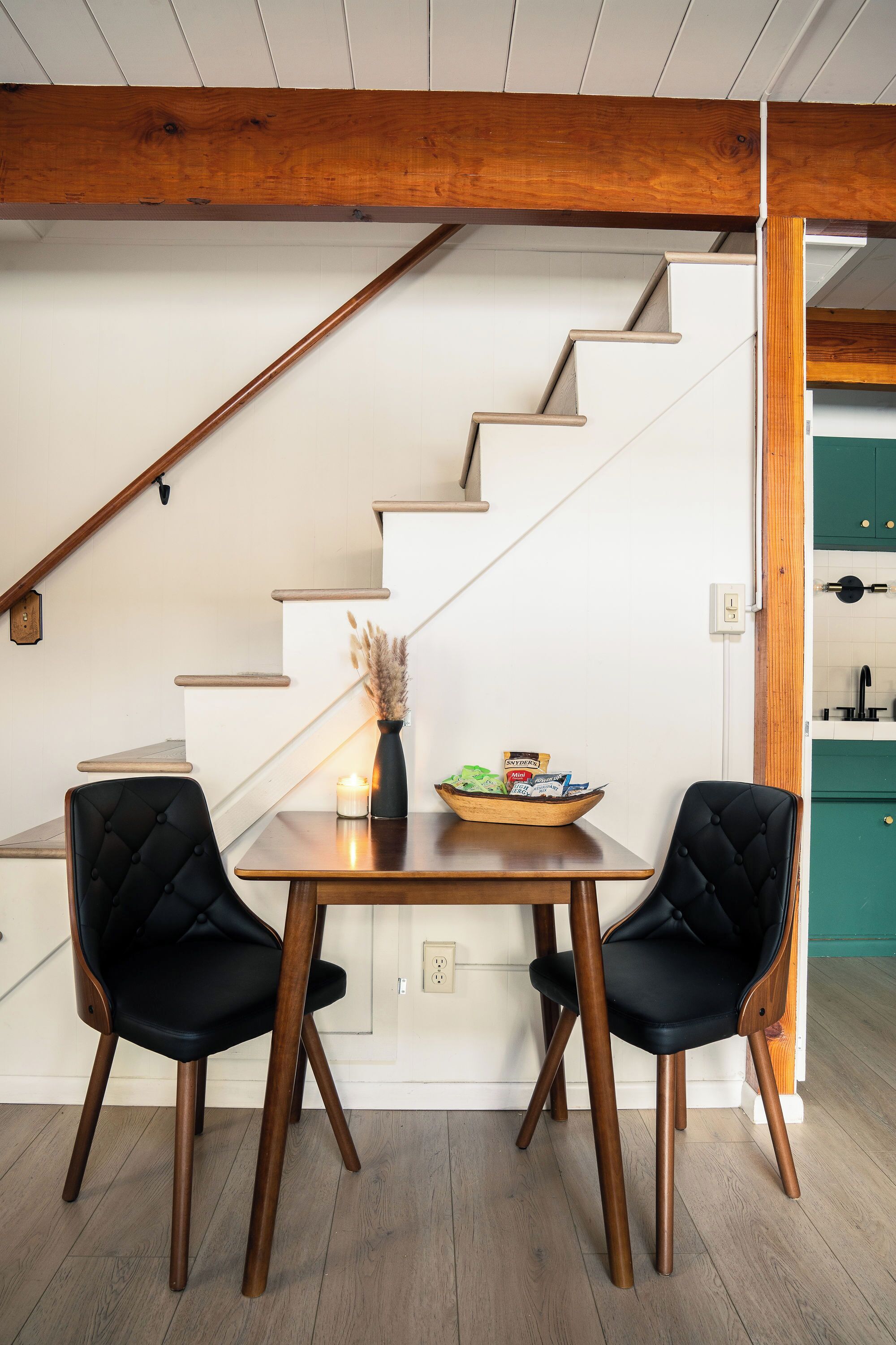 A dining area with a wooden table, two black upholstered chairs, a vase with dried plants, and a bowl of snacks.