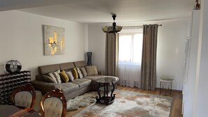 A living room with a sectional sofa, a dining table with chairs, a chandelier, and a large window with curtains.