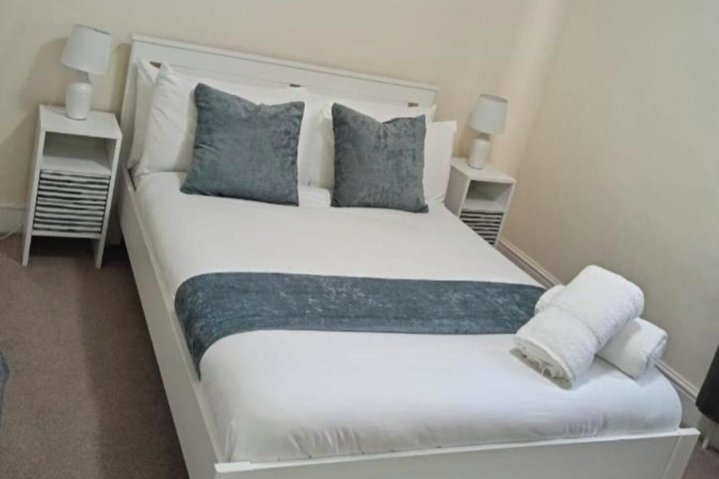 A neatly made bed with a white and grey duvet, two pillows, and a grey throw blanket. There are two bedside tables with lamps and a small shelf on each.