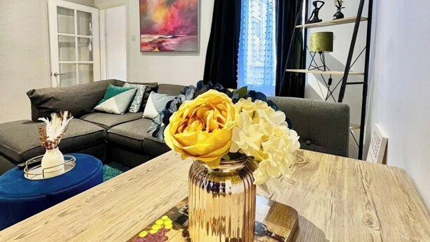 Ein Wohnzimmer mit Couch in Grau, einem blauen Ottomanen, einem Holztisch und einer Vase mit Blumen.
