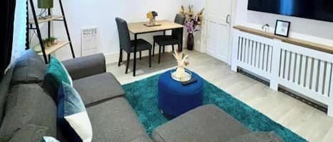 Ett vardagsrum med en hörnsoffa, ett matbord med stolar, en tv som är monterad på väggen och en liten växt på fönsterbrädan.