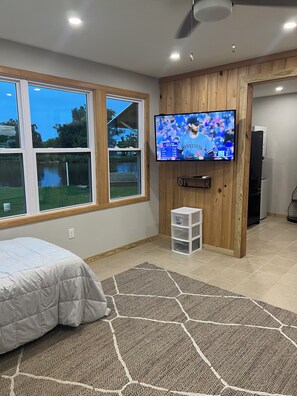 Quarto com cama, televisão fixada numa parede de madeira e vista para um lago através de amplas janelas.
