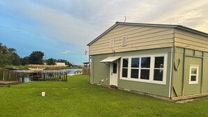 A small, single-story building with a green exterior and white trim. It features a large window on the front and a door with a small porch area.