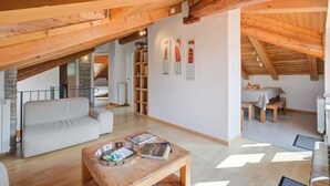 A spacious loft with a high ceiling, wooden beams, a white sofa, a wooden coffee table with books, and a dining area with a table and chairs.