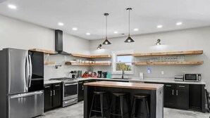 A modern kitchen with a central island, bar stools, stainless steel appliances, and wooden shelves.