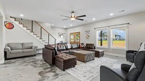 A spacious living room with a staircase, a large sectional sofa, a coffee table, and a view of the outdoors through large windows.
