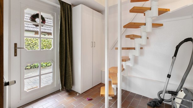 Een kamer met een witte deur, een raam met een krans, een trap met houten treden en een stofzuiger.