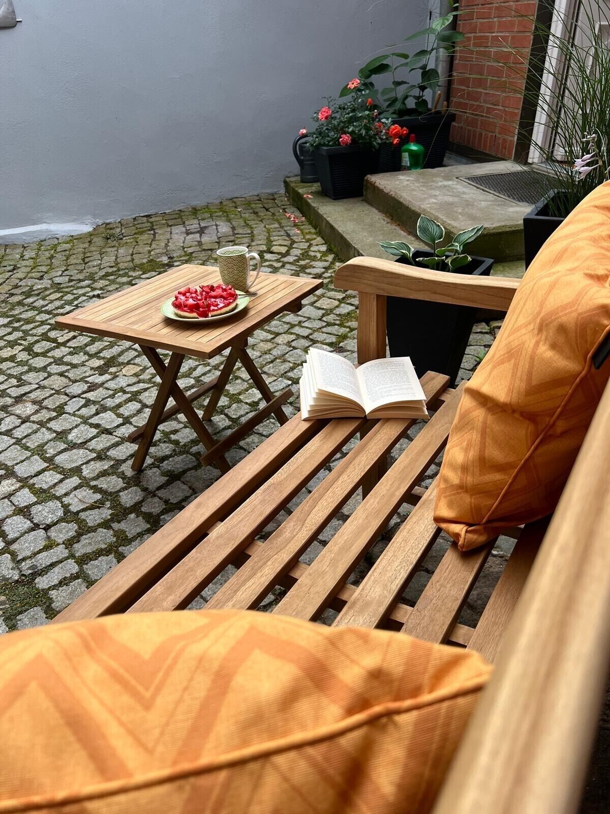 Eine Holzbank mit Kissen, ein kleiner Tisch mit Essen und einem Buch, sowie Blumentöpfe in einem Innenhof.