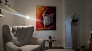 Moderni olohuone, jossa on harmaa sohva, pieni pyöreä pöytä, jonka päällä on sininen esine, tanssijaa esittävä maalaus ja kirja, joka on asetettu jalkarahin päälle.