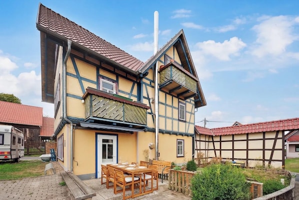 Une maison traditionnelle à colombages, dotée d’un balcon, d’un coin repas avec une table et des chaises, et d’un petit jardin.