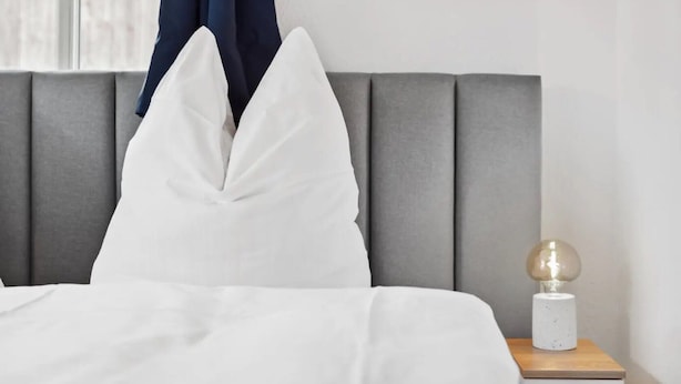 Una cama bien hecha con ropa blanca, un cabecero tapizado en gris y una mesita de noche con una lámpara.