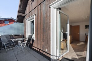 A modern outdoor patio with a glass door leading to a room, a wooden exterior wall, and a clear view of the adjacent building.