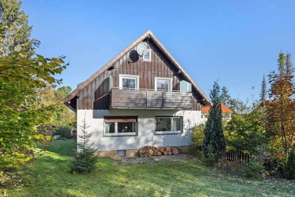 A two-story house with a sloped roof, a balcony, and a small front yard with a stack of firewood.