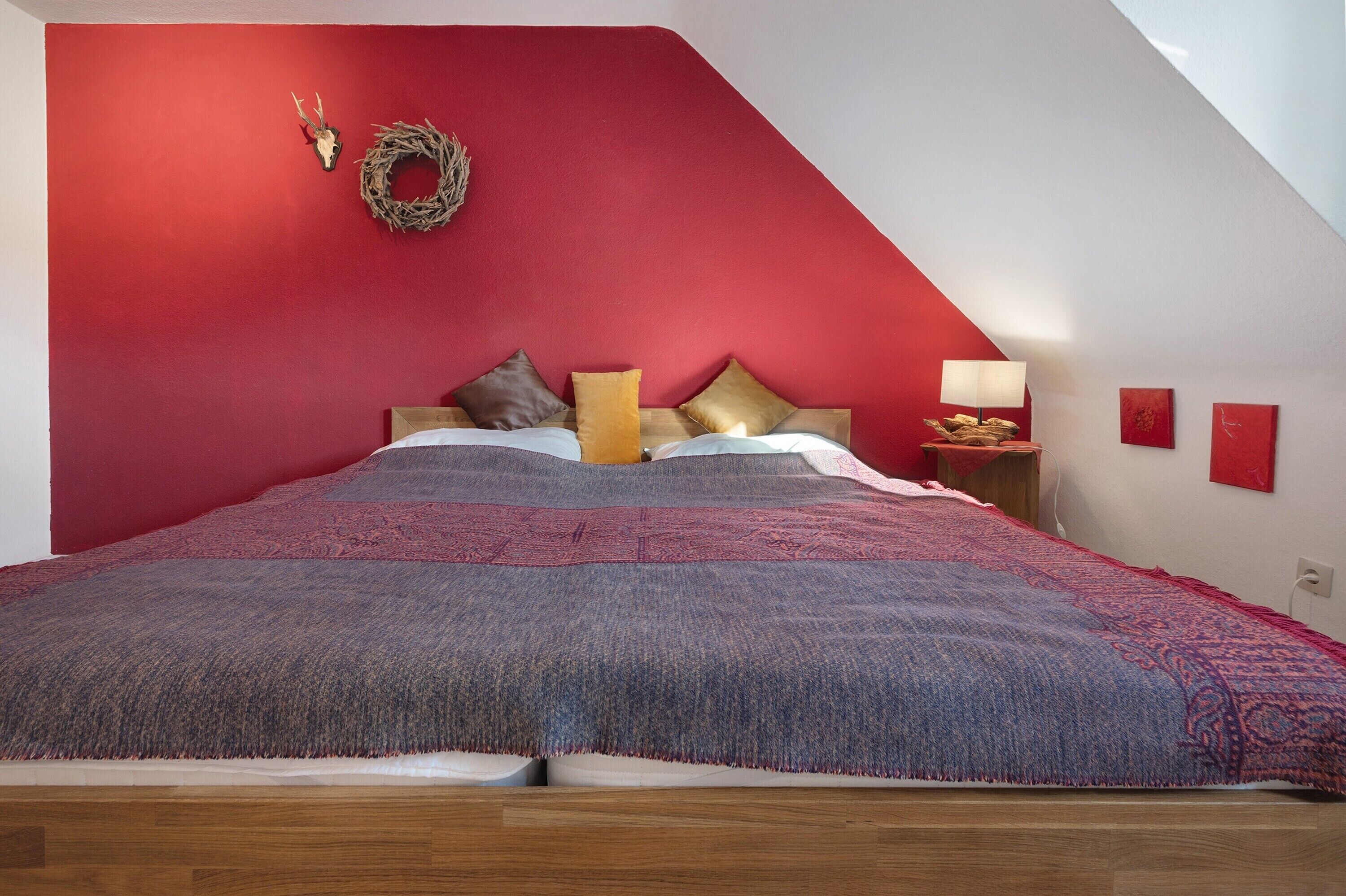 A bedroom with a red accent wall, a bed with patterned bedding, a nightstand with a lamp, and a wall-mounted wreath.