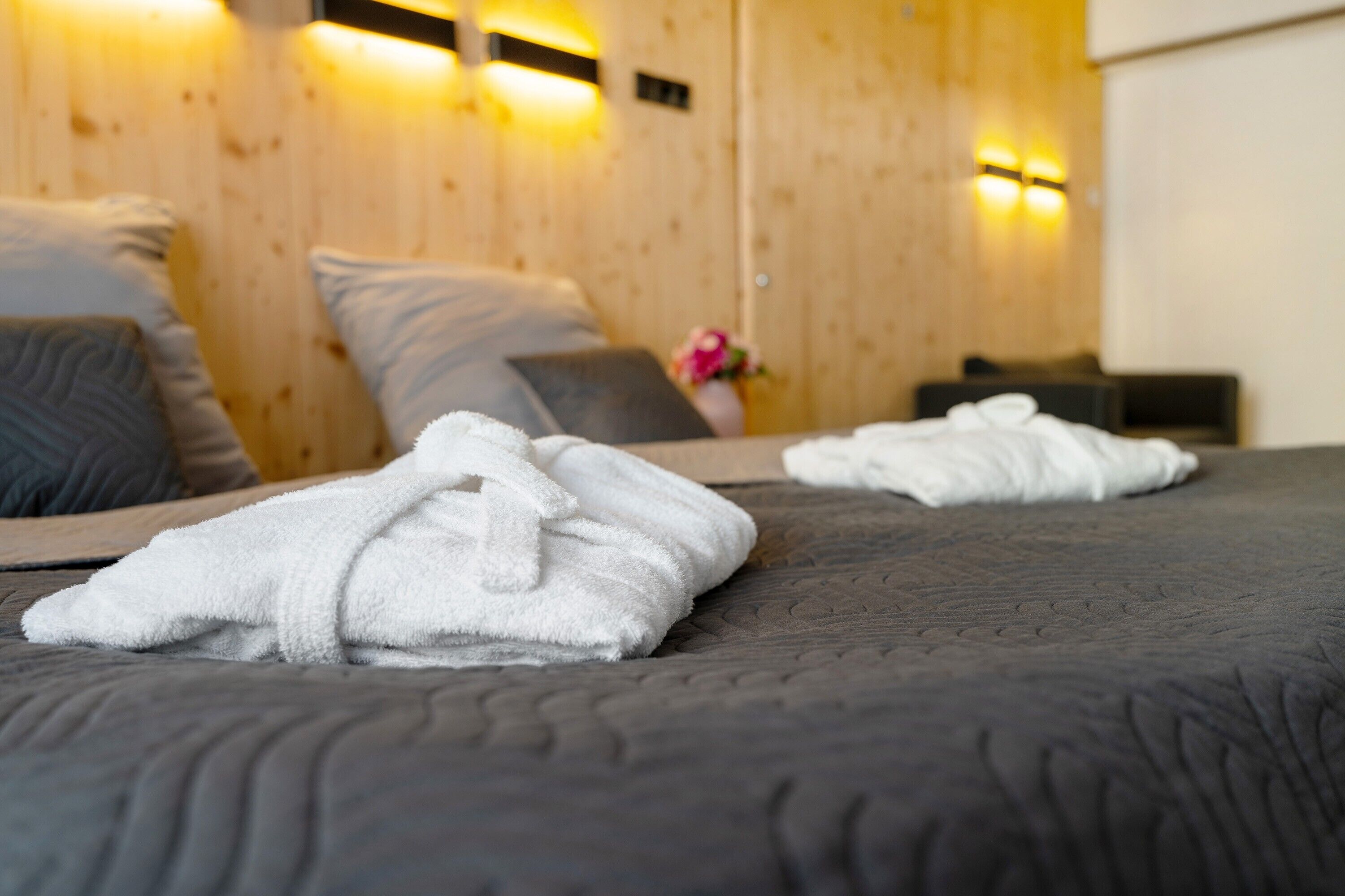 A bed with a dark quilt, a white towel, and a wooden wall with wall-mounted lights.