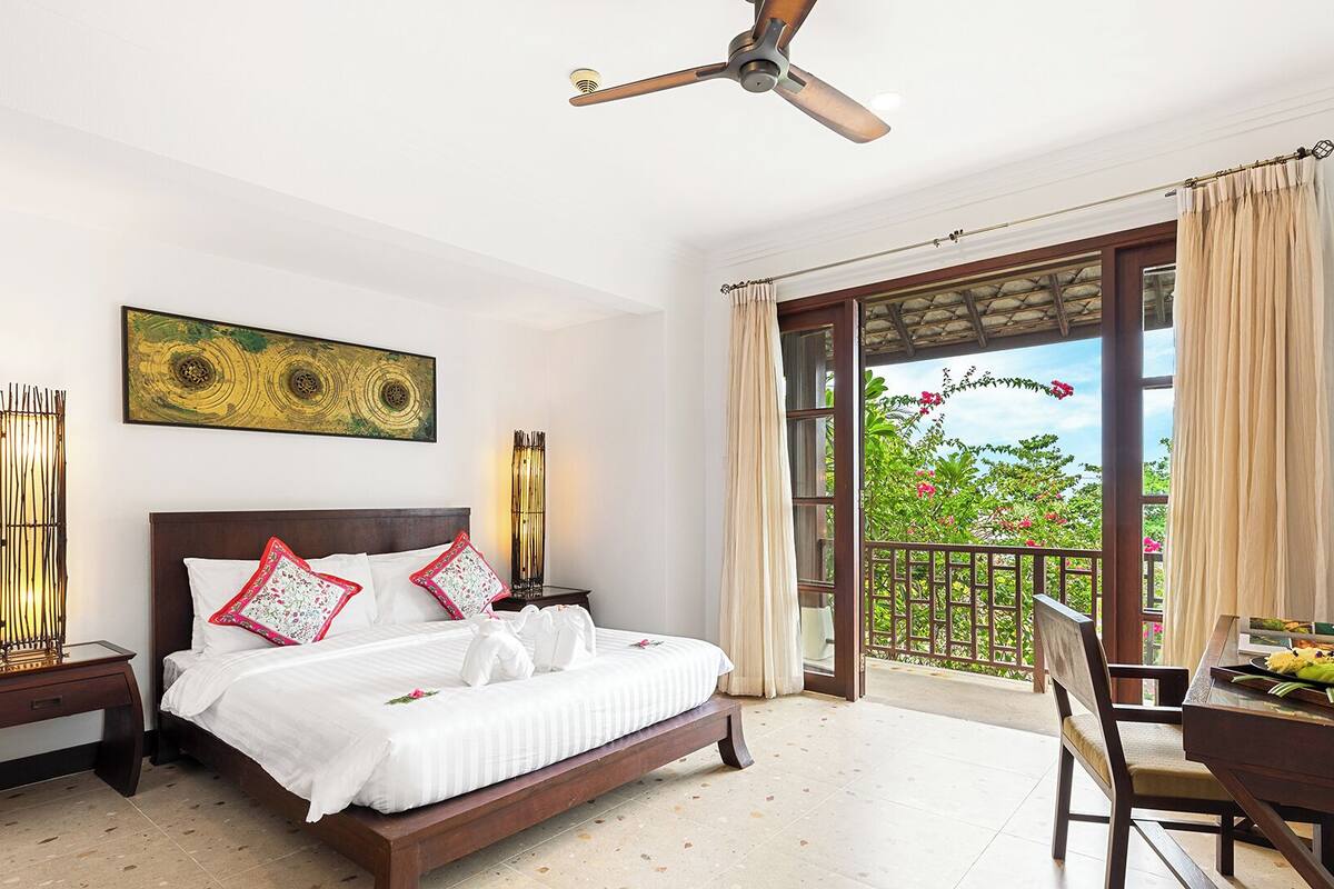 A bedroom with a bed, a ceiling fan, a dining table with a chair, and a balcony with a view of greenery.