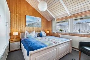 A bedroom with a wooden ceiling, a bed with a blue bedspread, two bedside lamps, a window with a view, and a radiator. - Holiday Home 'Haus Albertsen' with Private Terrace, Private Garden and Wi-Fi (Hellschen-Heringsand-Unterschaar)