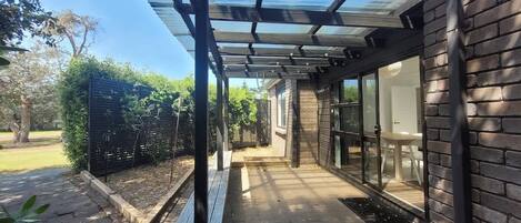A covered patio area with a bench, a glass door leading to a room, and a brick wall.