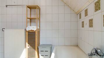 Ein Badezimmer mit einer Badewanne, einem hölzernen Ablagebrett mit Handtüchern, einem Heizkörper und gemusterten Fliesen an der Wand.