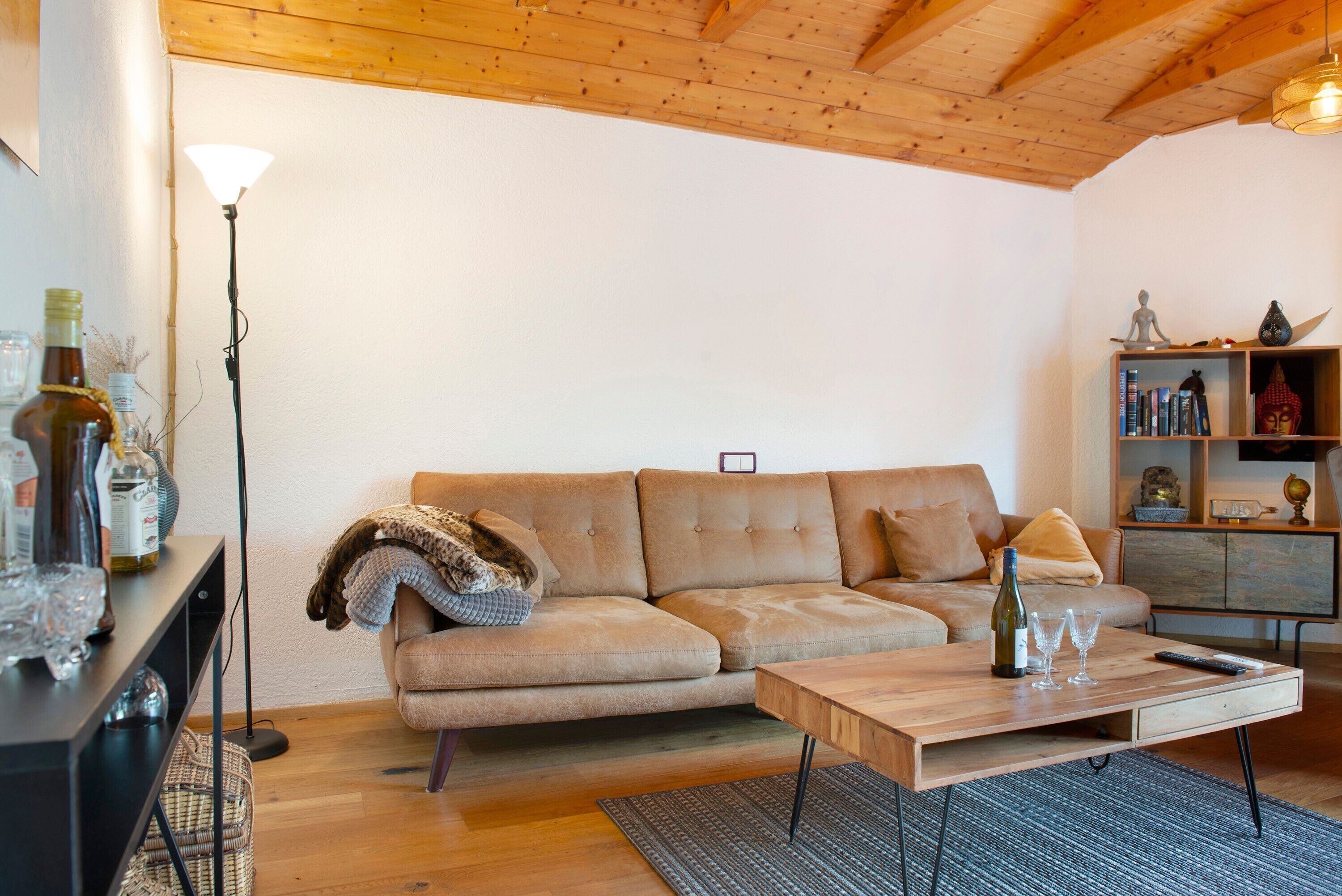 A living room with a sofa, a coffee table, a lamp, a bookshelf, and a wooden ceiling.