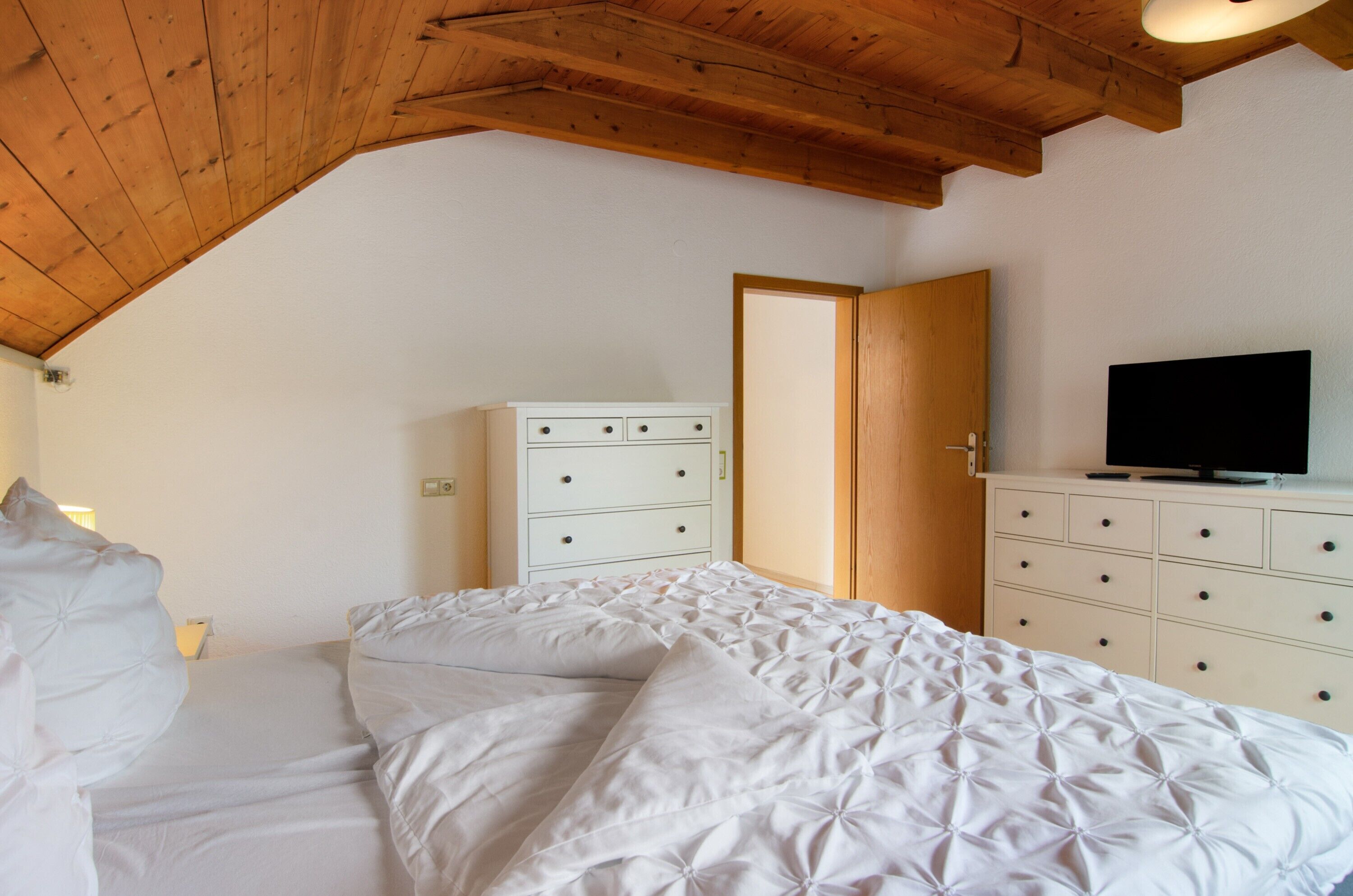 A bedroom with a wooden ceiling, a bed with white bedding, a dresser with drawers, and a television on top.