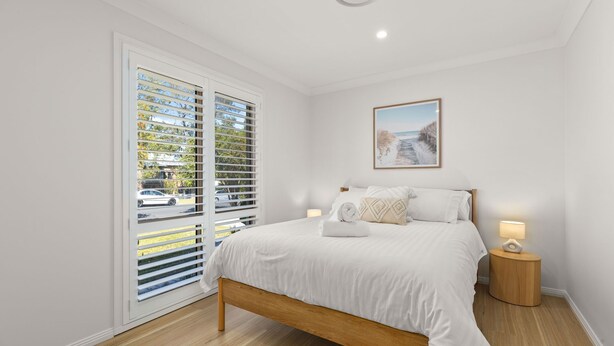 A bedroom with a bed, a nightstand, a lamp, a window with blinds, and a framed picture on the wall.
