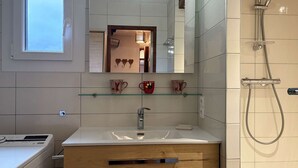 A modern bathroom with a large mirror, a shower, a sink with a wooden cabinet, and a window.