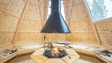 A wooden cabin with a central fireplace, benches, and a large window.