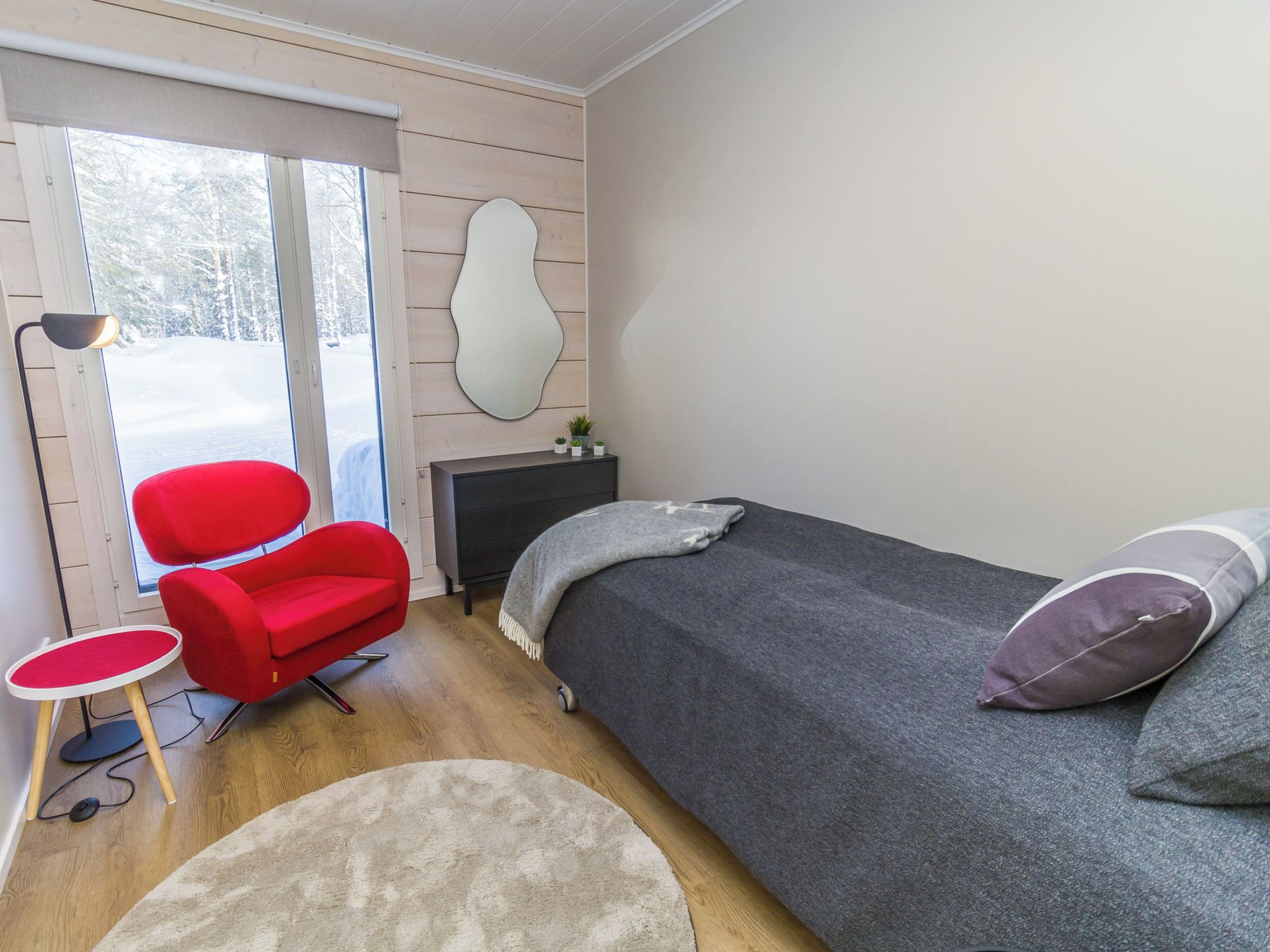 A bedroom with a bed, a red chair, a small table, a lamp, and a window with a view of trees.