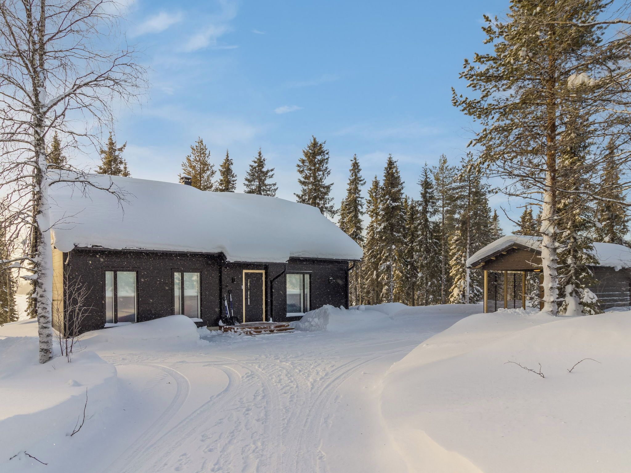 Lumihunnun peittämä mökki, jonka ympärillä on puita ja lumipeitteinen maisema.