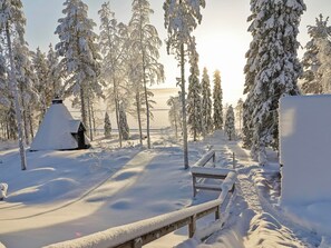 A snowy landscape with a wooden bench and a snow-covered structure, likely a cabin or hut, surrounded by trees.