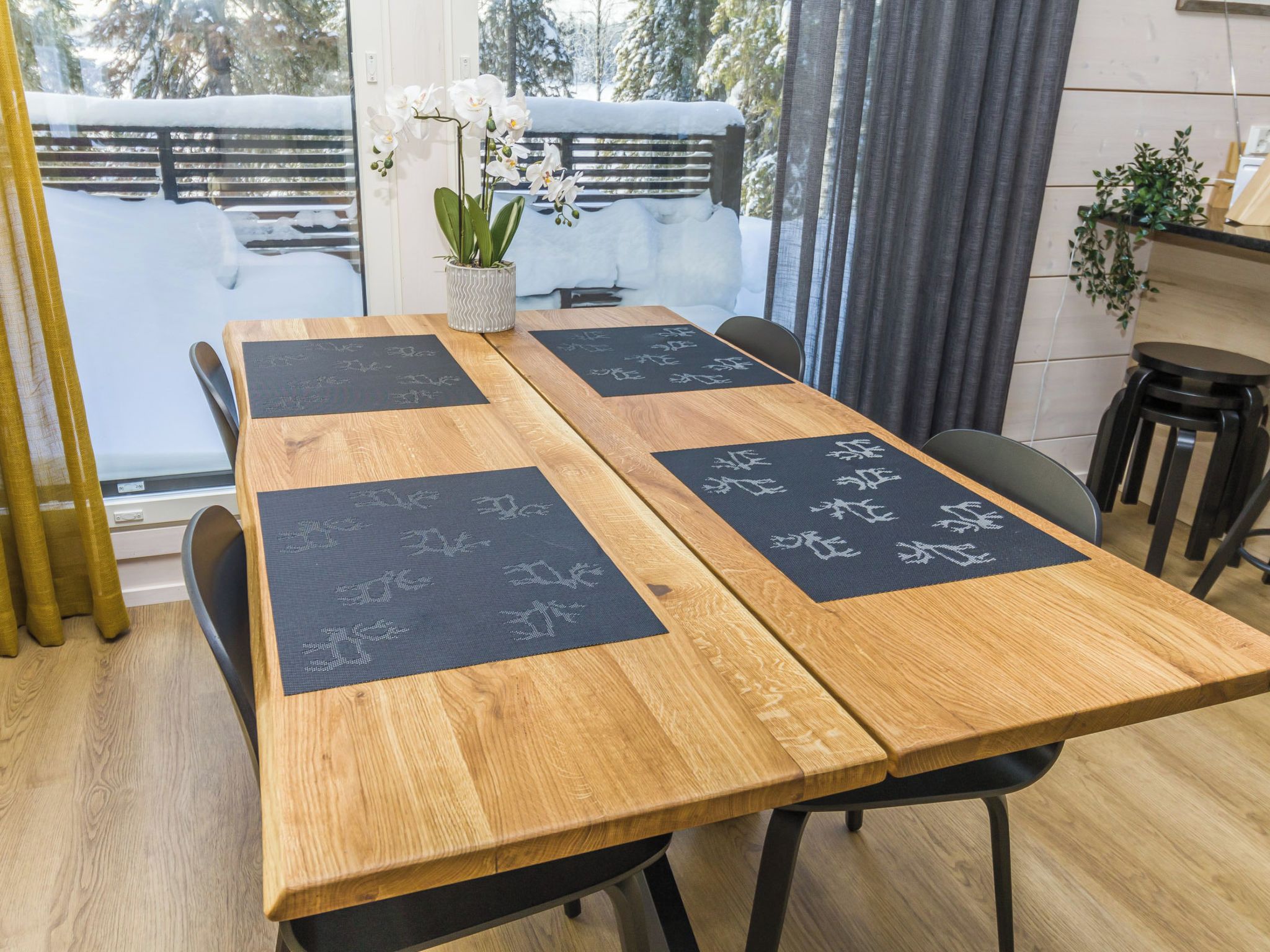 Een houten eettafel met zwarte placemats met witte boommotieven, omringd door stoelen, met een potorkidee en uitzicht op een besneeuwd landschap.