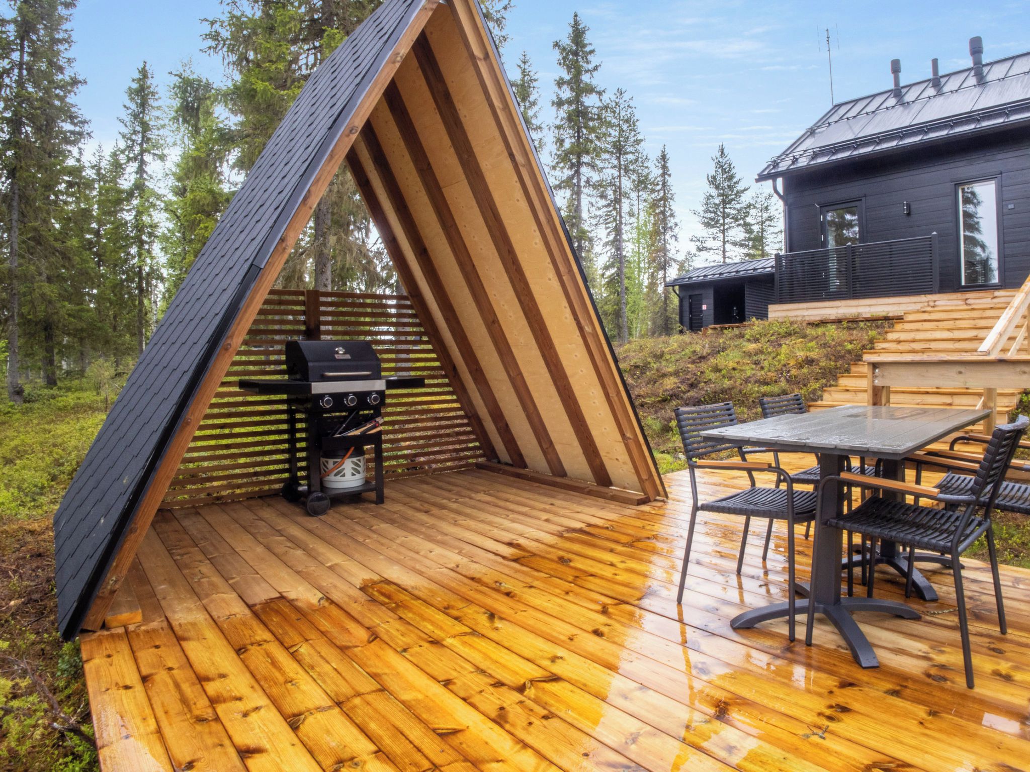 Een terras van hout buiten met een barbecue, een tafel met stoelen en een huis op de achtergrond.