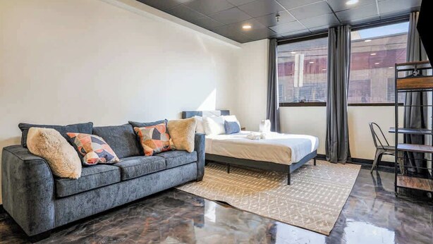 Een moderne hotelkamer met een grijze bank, een bed, een klein bureau en een groot raam.