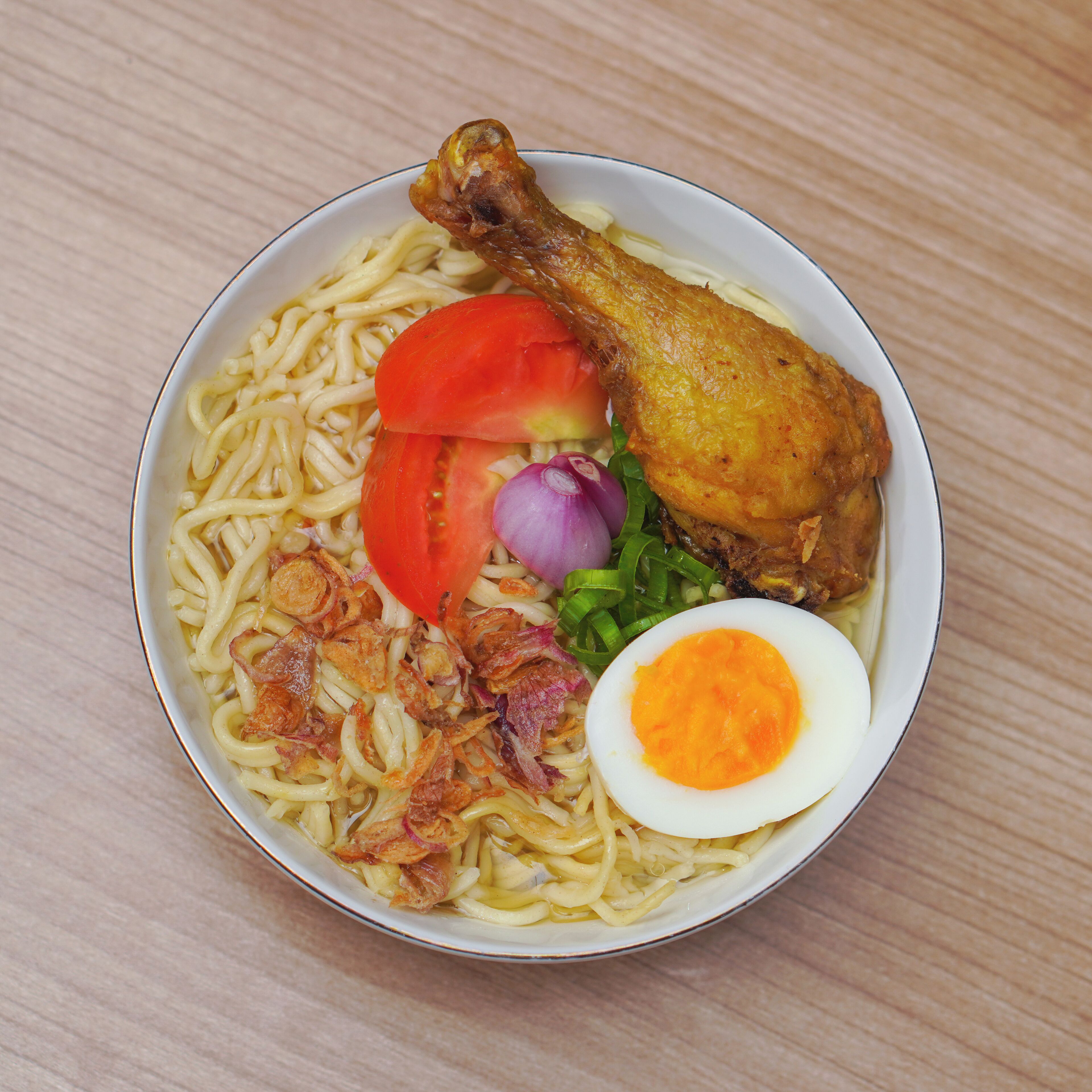 A bowl of noodles with a fried chicken leg, a slice of tomato, a boiled egg, and some green onions.