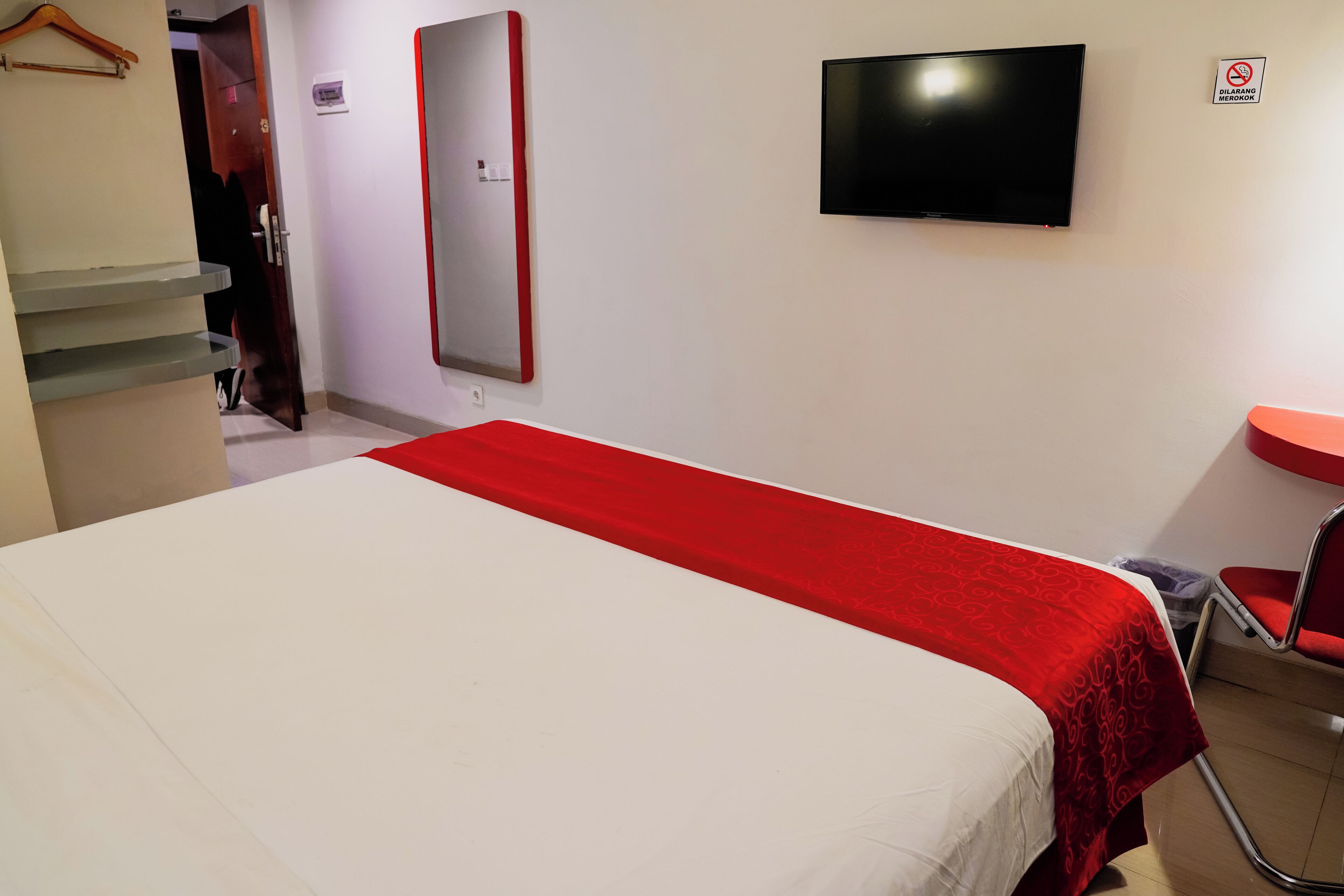 Habitación de hotel con cama, televisor, una silla roja, una mesita y un espejo.