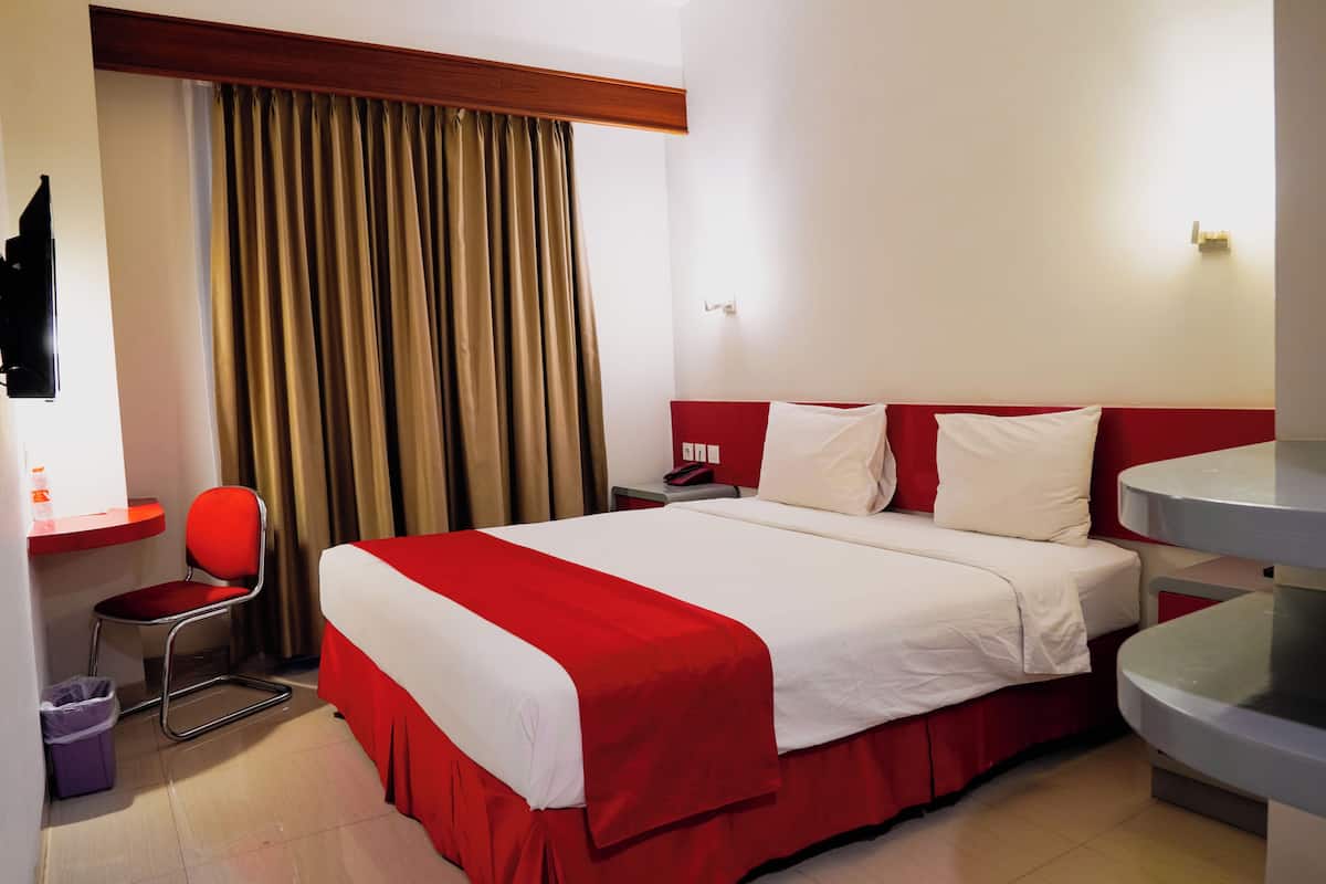 A hotel room with a bed, a chair, a desk, a trash bin, and a window with curtains.