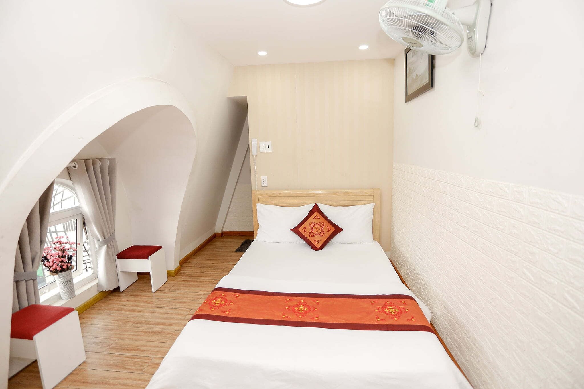 A hotel room with a bed, a small table, a chair, a window with curtains, and a wall-mounted fan.