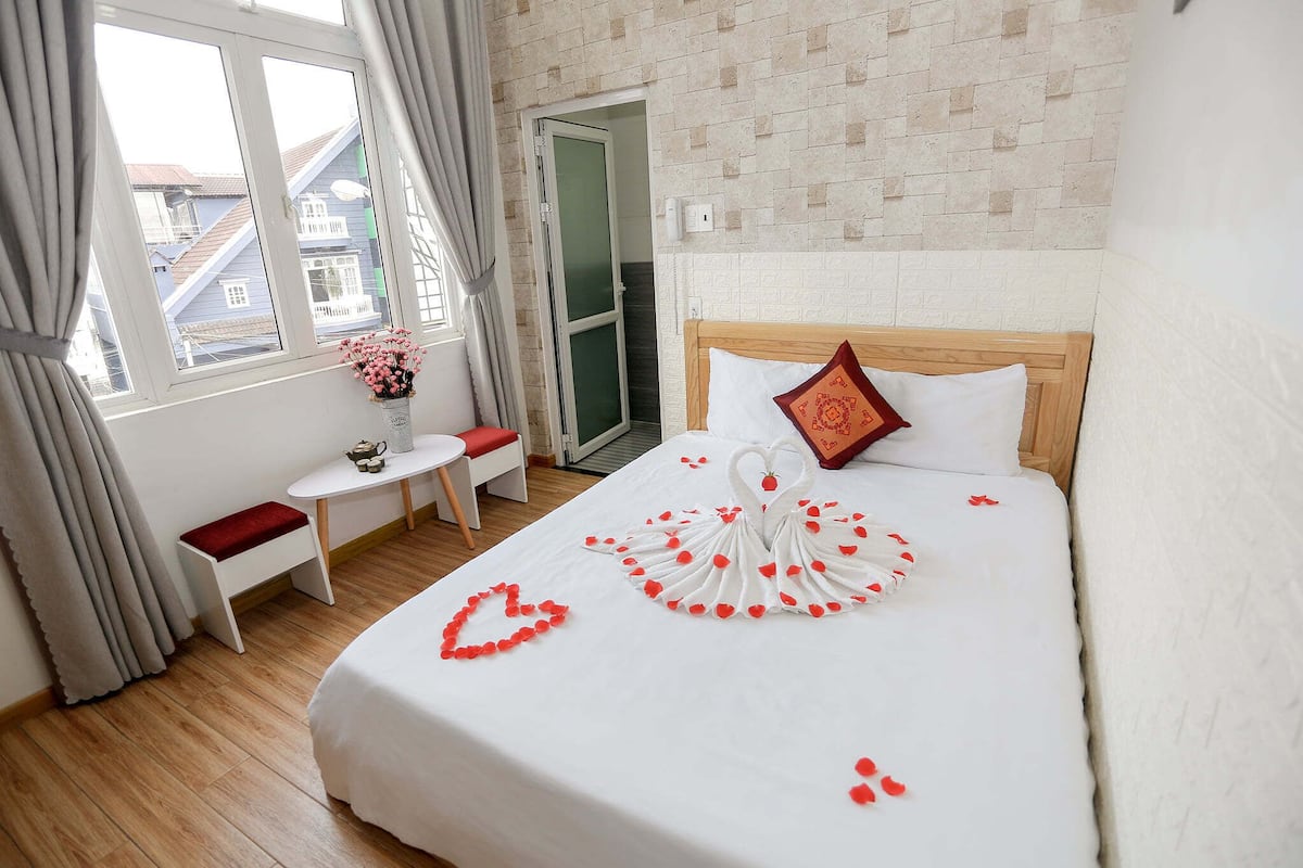 Une chambre d’hôtel avec un lit, une petite table et une chaise, une fenêtre avec des rideaux et une vue sur les bâtiments environnants.