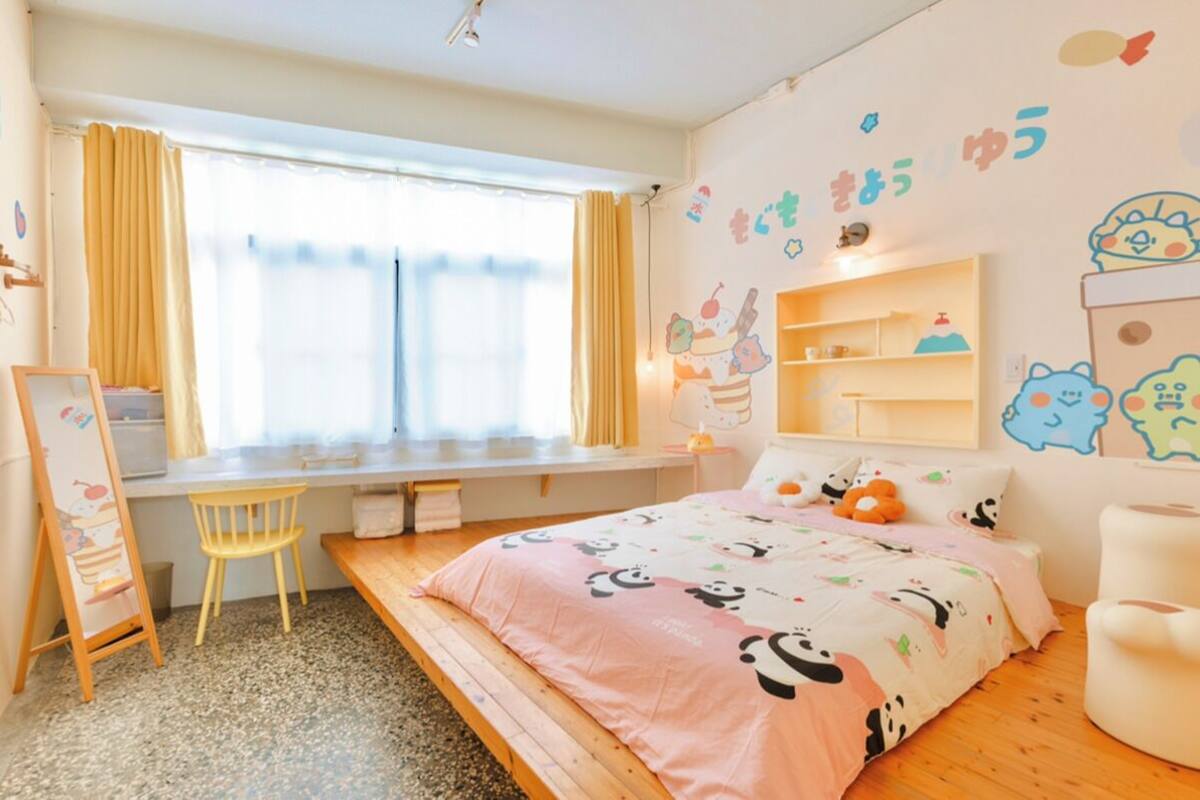 A child-friendly bedroom with a bed, desk, and chair, featuring playful wall decorations and a window with curtains.
