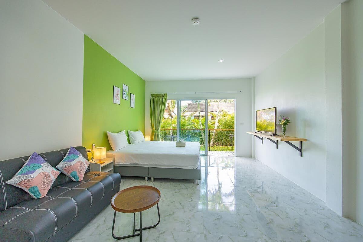 A modern hotel room with a green accent wall, a bed, a sofa, a small table, and a TV mounted on the wall.